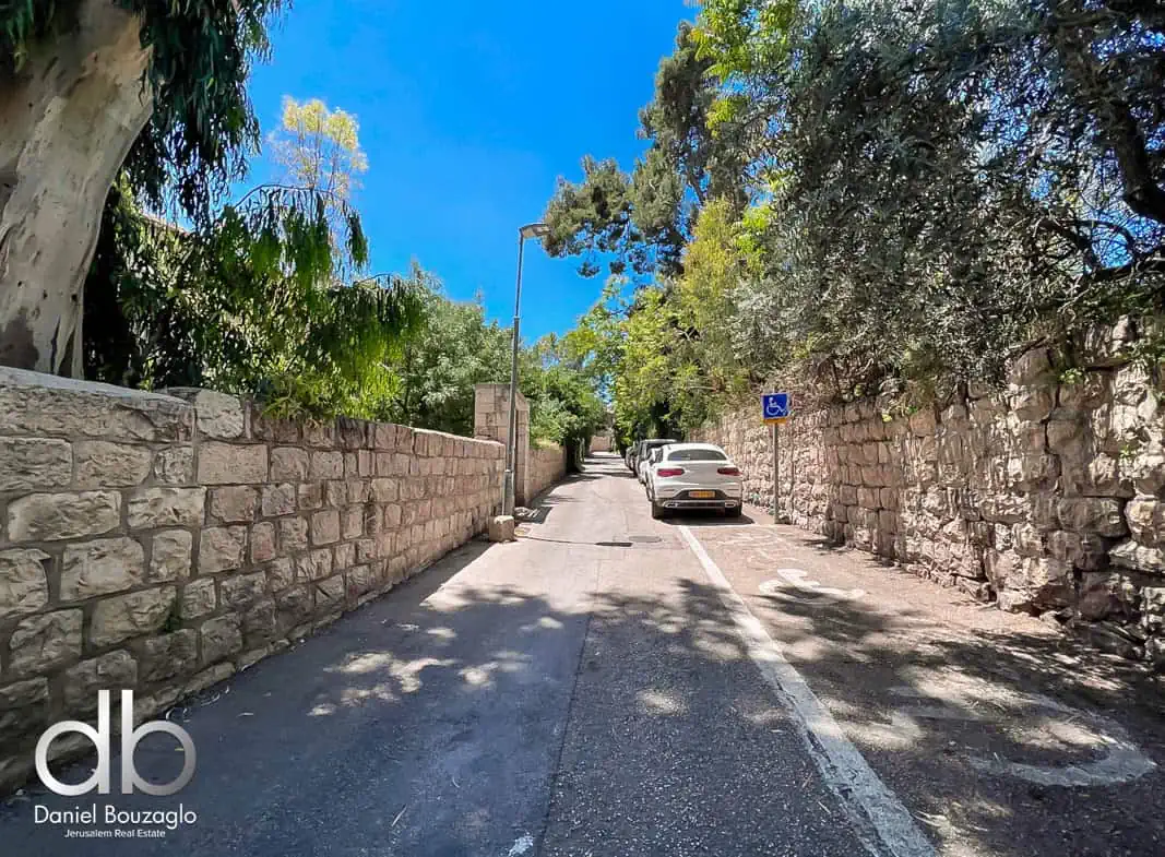Sale sign for Jerusalem real estate property by Daniel Bouzaglo, on a quiet residential street with parked cars and lush greenery.