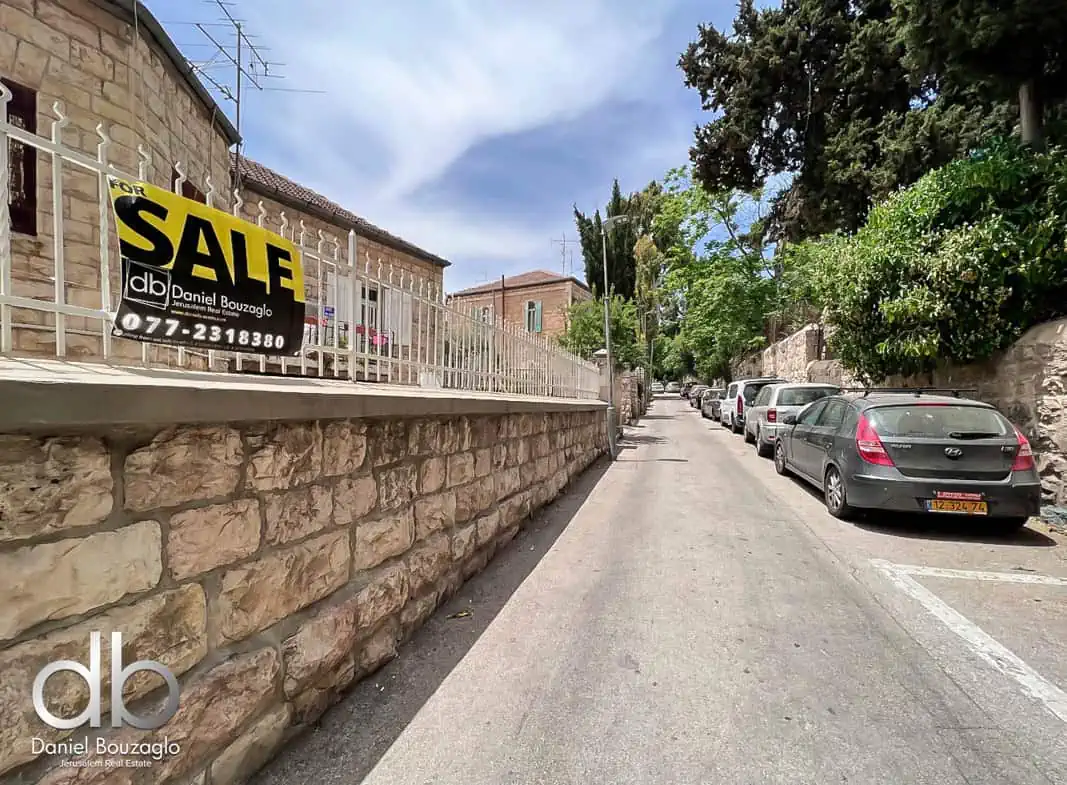 Sale sign for Jerusalem real estate property by Daniel Bouzaglo, on a quiet residential street with parked cars and lush greenery.