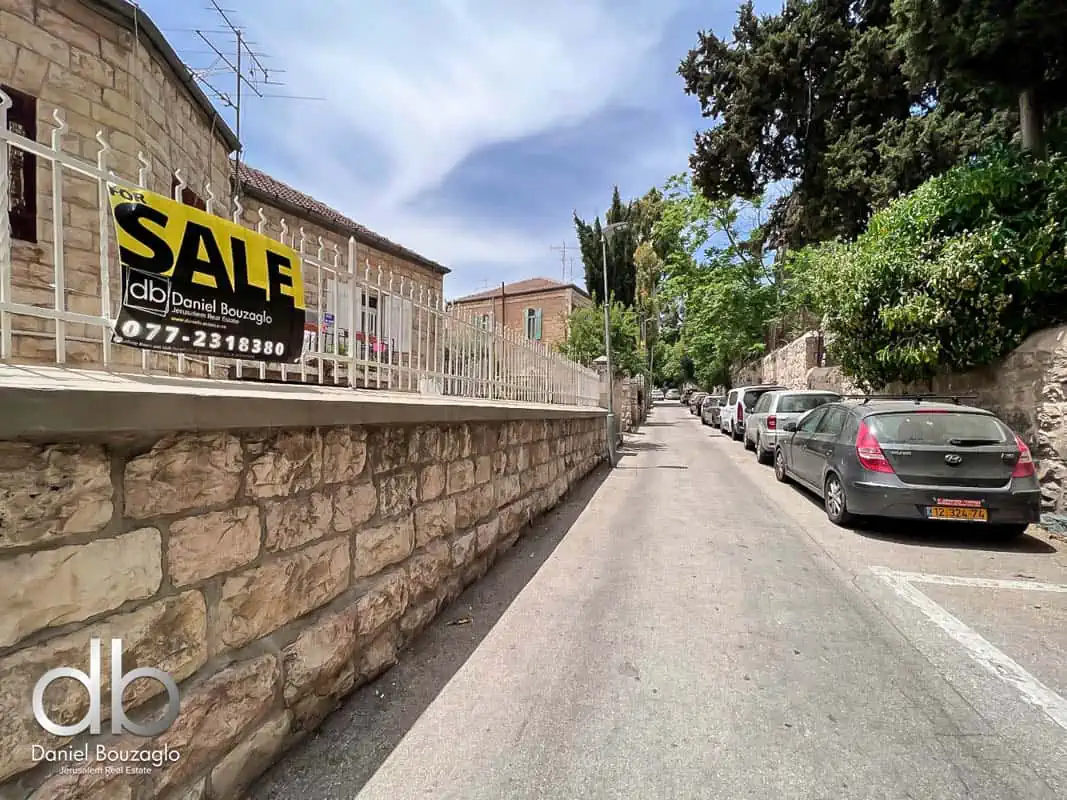 Sale sign for Jerusalem real estate property by Daniel Bouzaglo, on a quiet residential street with parked cars and lush greenery.