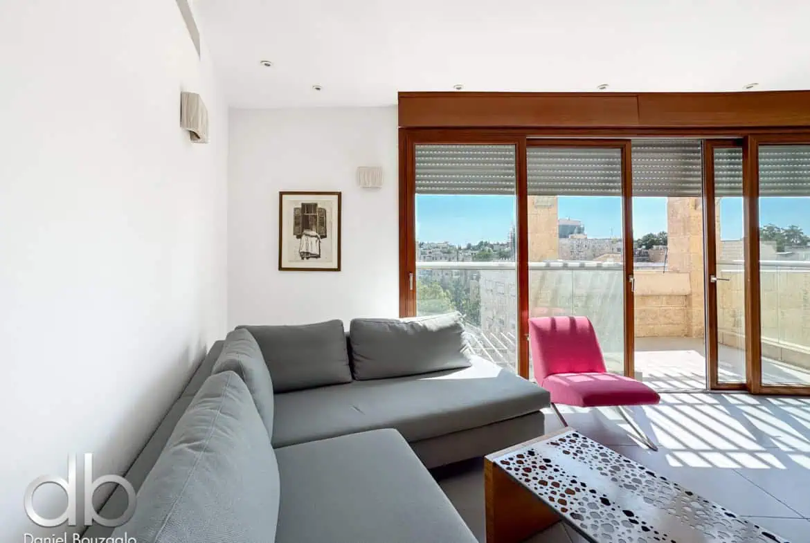David Crown 25br furnished rental Jerusalem 12 Modern Jerusalem apartment living room with a spacious balcony, large glass sliding doors, stylish grey sofa, and bright pink accent chair, showcasing Jerusalem real estate and interior design.