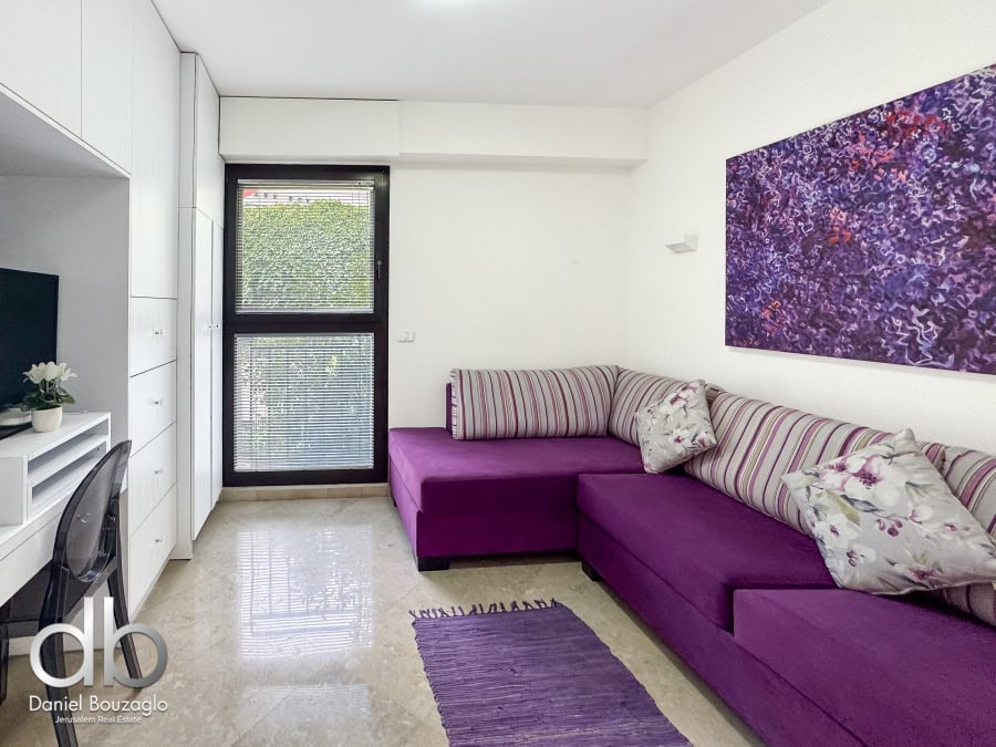 Garden apartment in David VIllage for sale 2 Bright Jerusalem apartment living room with modern furnishings and purple accents, featuring a L-shaped sofa, artwork, and large window for natural light.