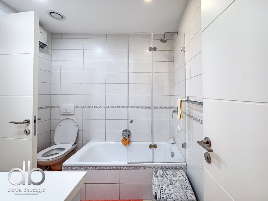 Garden apartment in David VIllage for sale 3 Modern Jerusalem real estate bathroom with white tiled walls, bathtub, and glass shower partition. Ideal for Jerusalem homes, featuring clean design and practical amenities.
