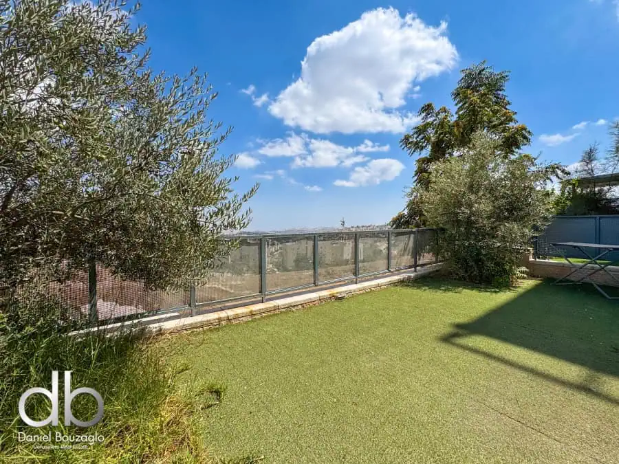 Balcony view with green artificial grass, lush trees, and city skyline in Jerusalem, Israel, under a bright blue sky with scattered clouds, showcasing Jerusalem real estate.