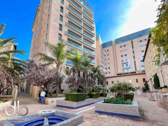 Courtyard of a modern beige apartment complex with palm trees, tiled planters, and a fountain in the foreground.