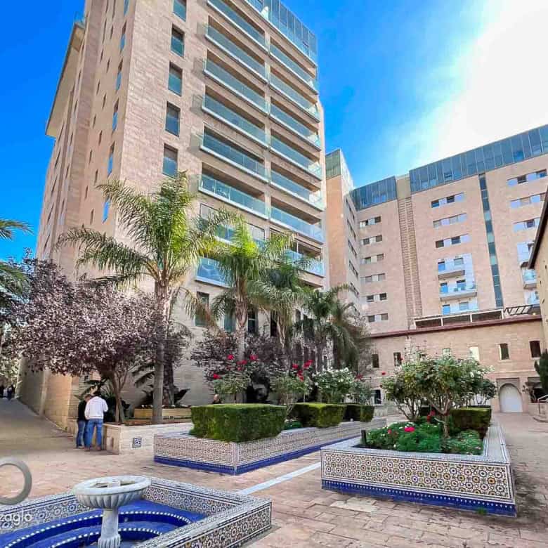 Courtyard of a modern beige apartment complex with palm trees, tiled planters, and a fountain in the foreground.