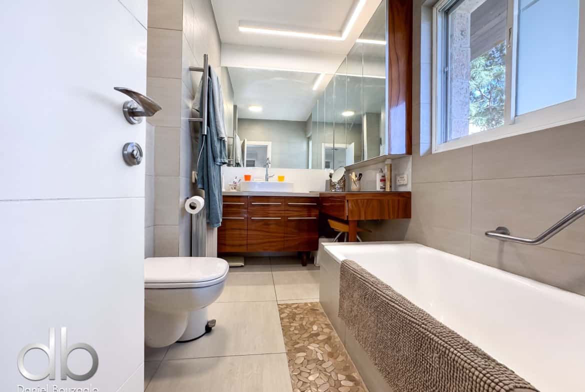 Light-filled modern bathroom with a wooden vanity, large mirror, and a white bathtub along the right, window showing greenery outside