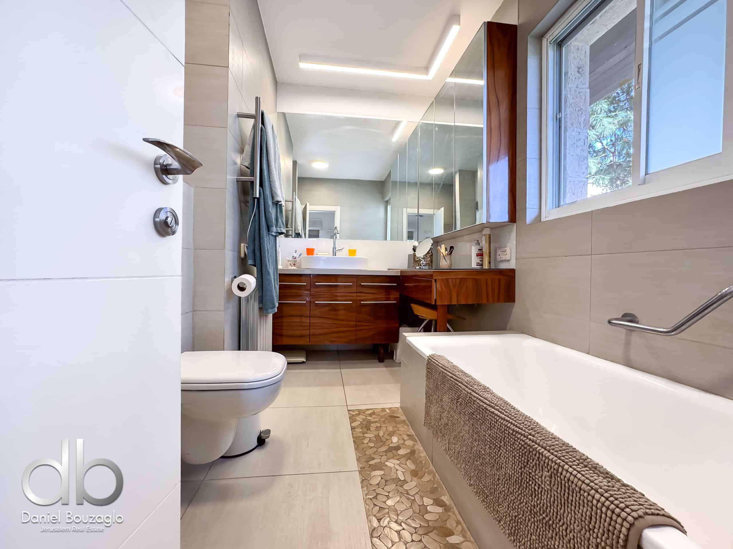 Light-filled modern bathroom with a wooden vanity, large mirror, and a white bathtub along the right, window showing greenery outside