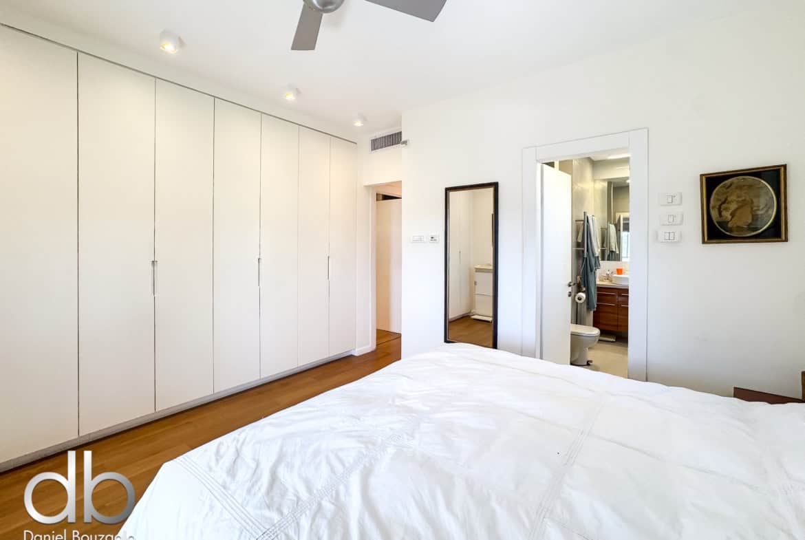 Bright modern bedroom with tall white built-in wardrobes along the left wall and a bed in the foreground; a doorway reveals a bathroom with a vanity mirror on the right center edge.