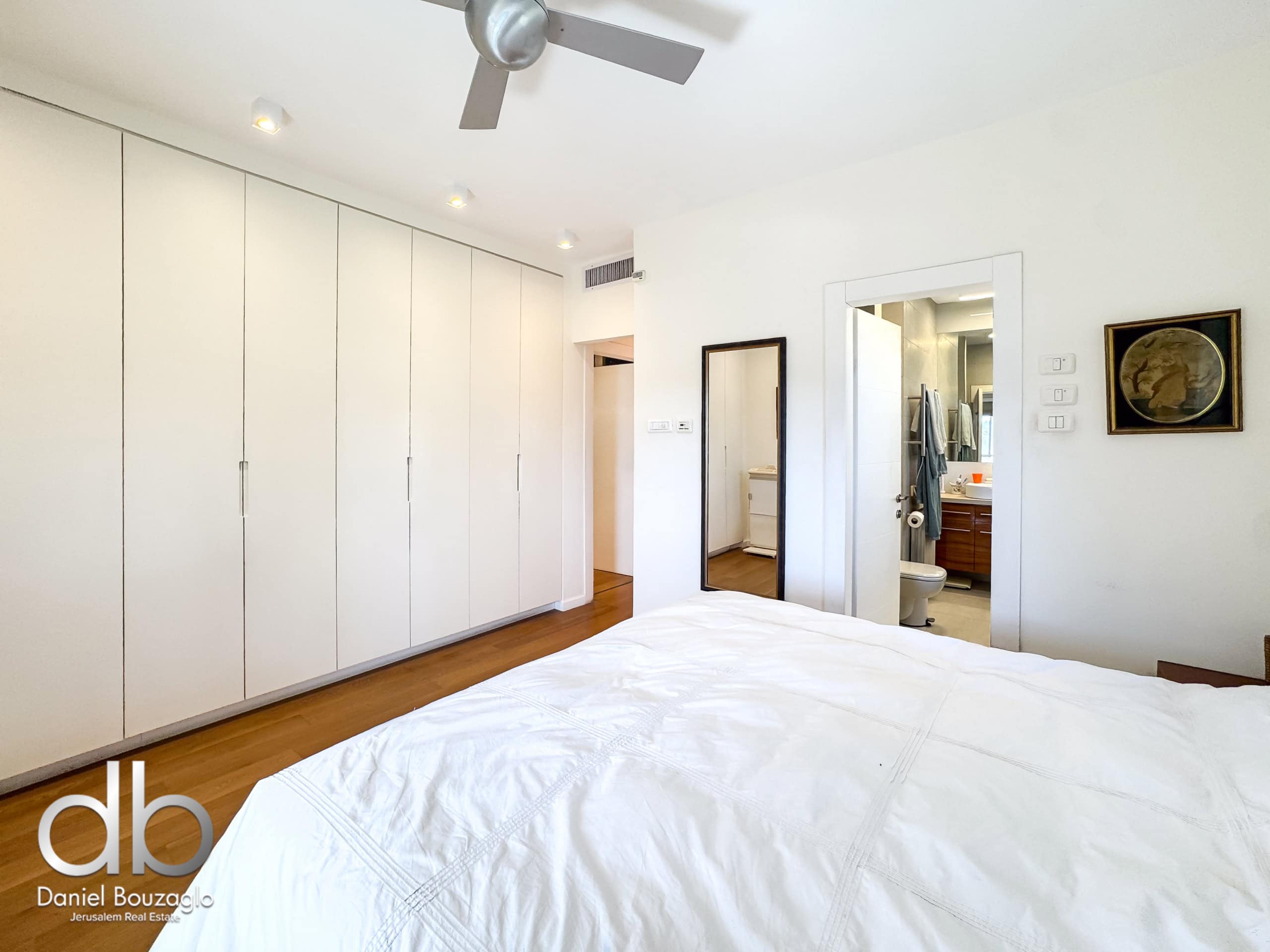 Bright modern bedroom with tall white built-in wardrobes along the left wall and a bed in the foreground; a doorway reveals a bathroom with a vanity mirror on the right center edge.