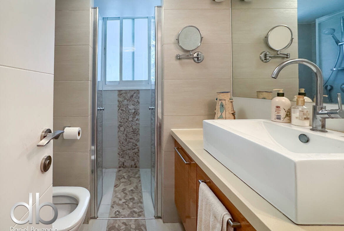 Modern bathroom showing a glass shower enclosure, a double-sink vanity, and beige tiled walls with round wall mirrors on either side of the sink area.