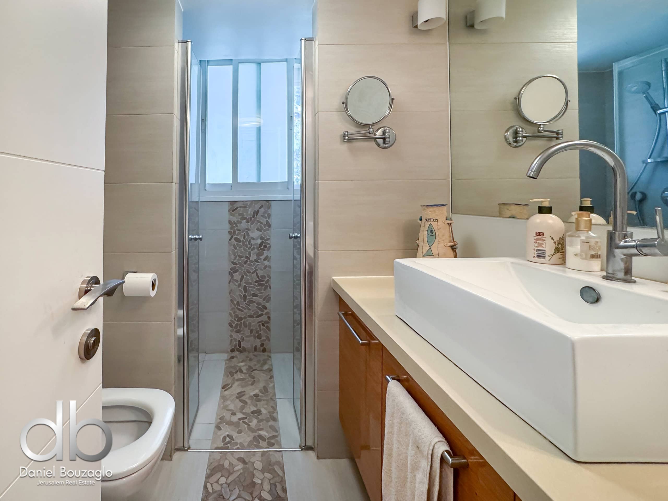 Modern bathroom showing a glass shower enclosure, a double-sink vanity, and beige tiled walls with round wall mirrors on either side of the sink area.