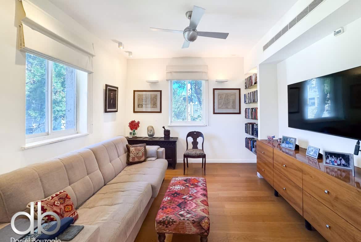 Bright living room with a beige tufted sofa, patterned red ottoman, and light wood floors; large windows and ceiling fan above.