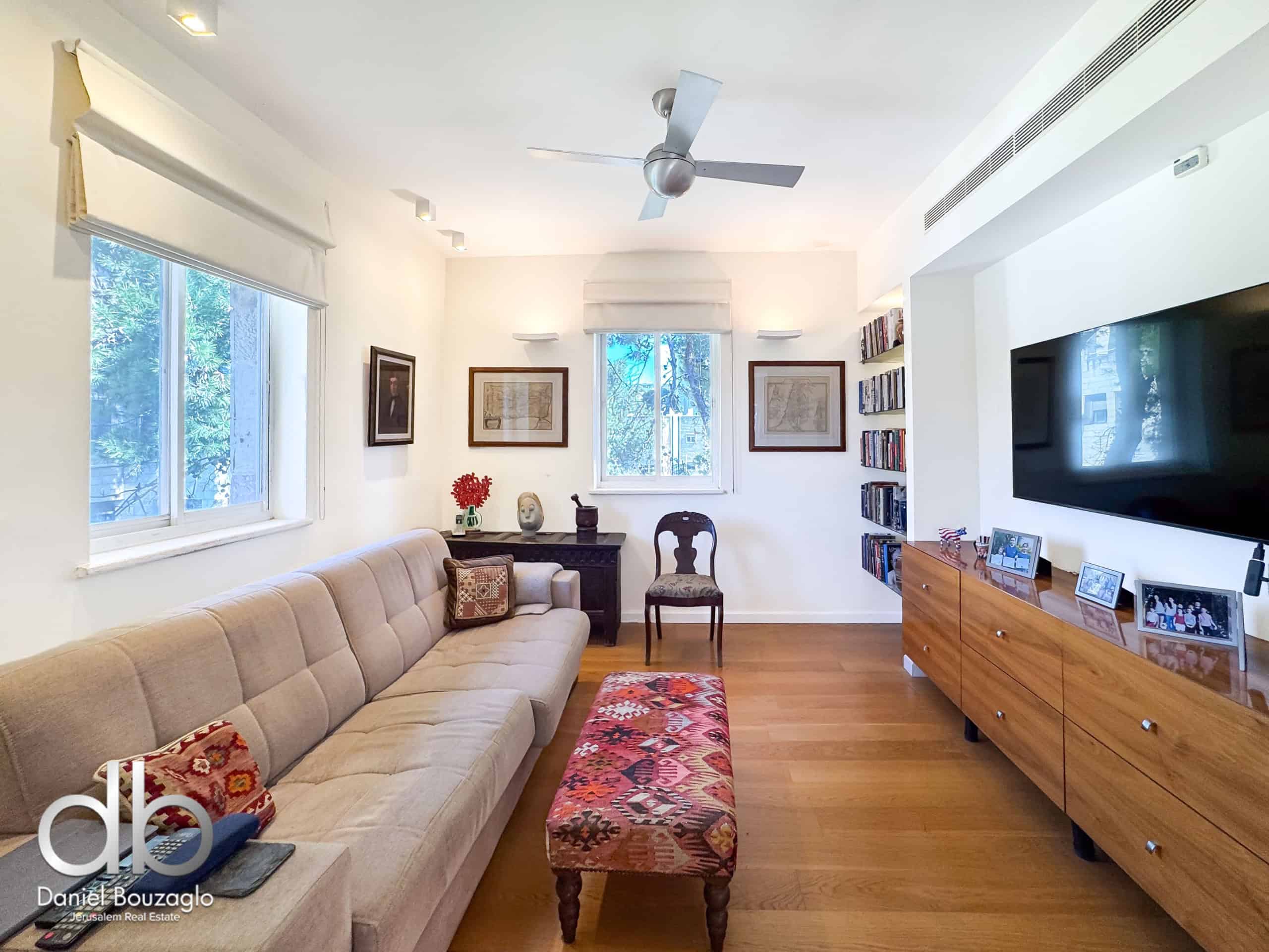 Bright living room with a beige tufted sofa, patterned red ottoman, and light wood floors; large windows and ceiling fan above.