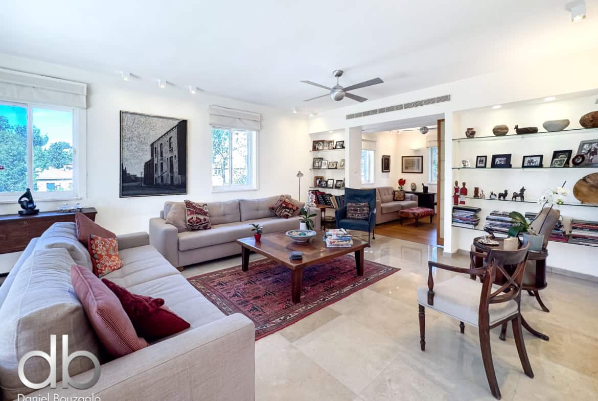 Bright contemporary living room with beige sectional sofa, red patterned throw pillows, and a wooden coffee table on a red rug; large windows bring in natural light, and built-in shelving displays photos and decor.