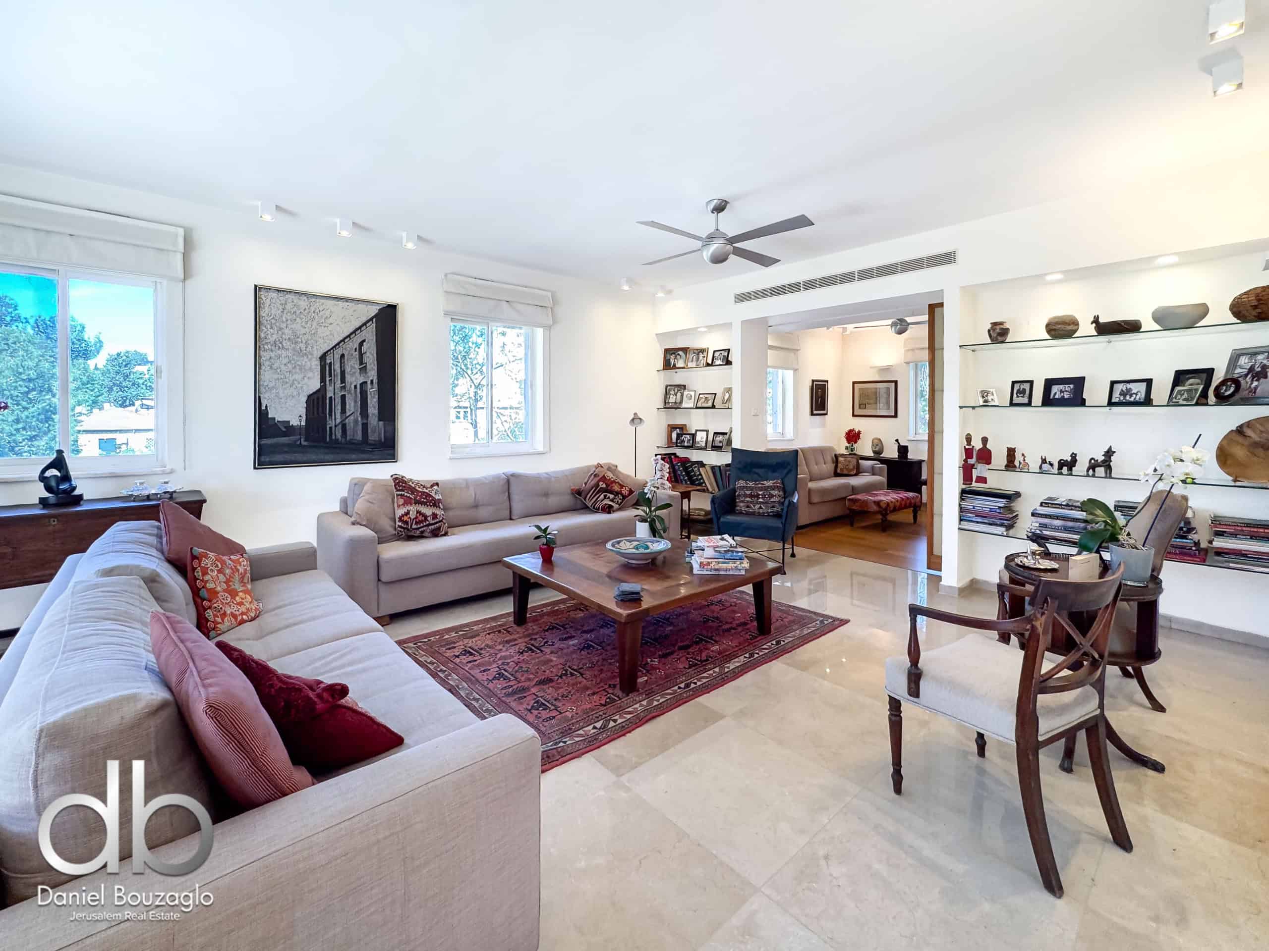 Bright contemporary living room with beige sectional sofa, red patterned throw pillows, and a wooden coffee table on a red rug; large windows bring in natural light, and built-in shelving displays photos and decor.