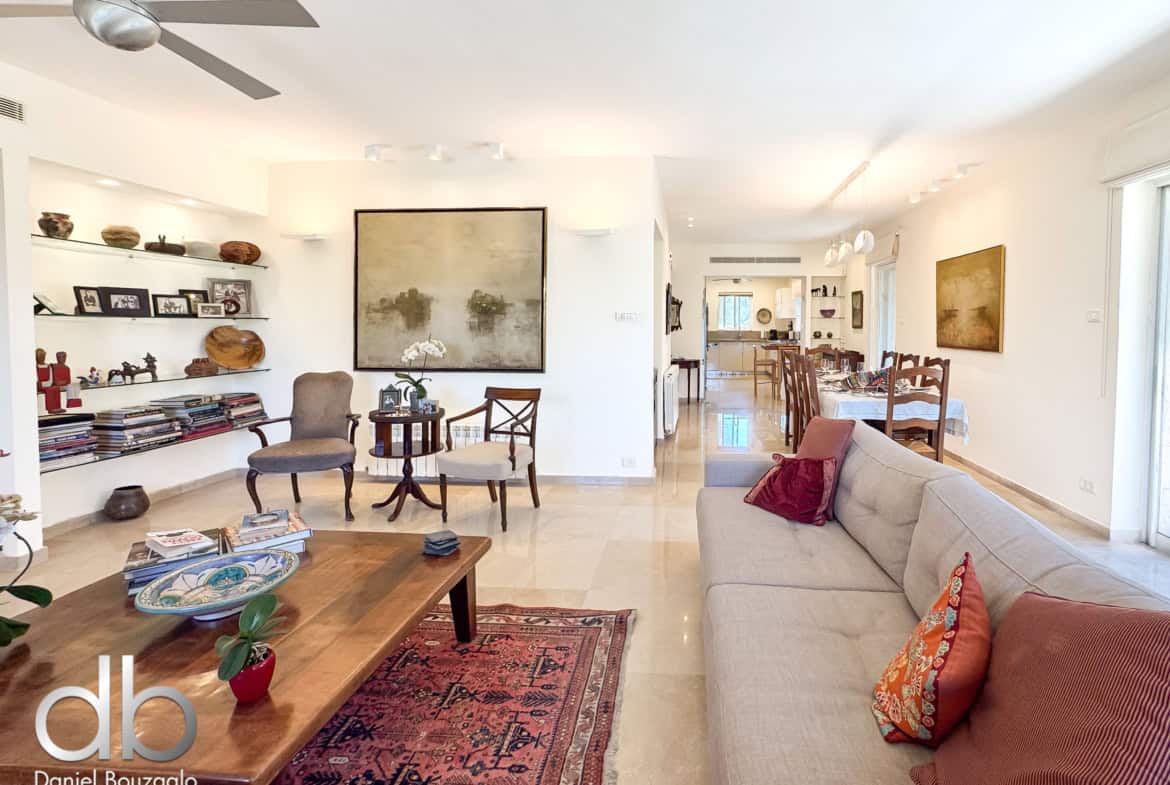 Open-plan living area with beige sofa set, wooden coffee table, and a red patterned rug; shelves with decor and photos line the wall, and the dining area is visible in the background.