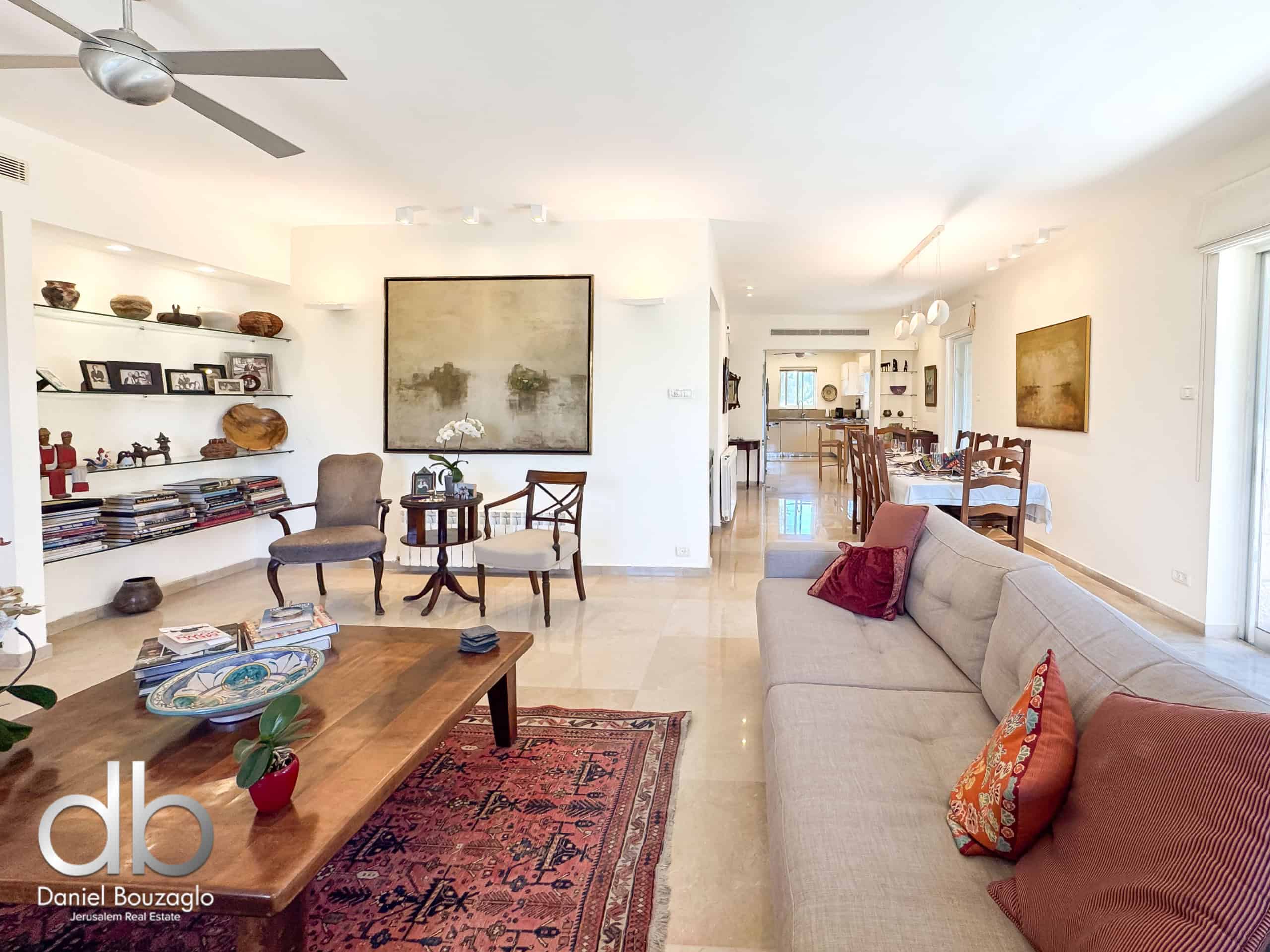 Open-plan living area with beige sofa set, wooden coffee table, and a red patterned rug; shelves with decor and photos line the wall, and the dining area is visible in the background.