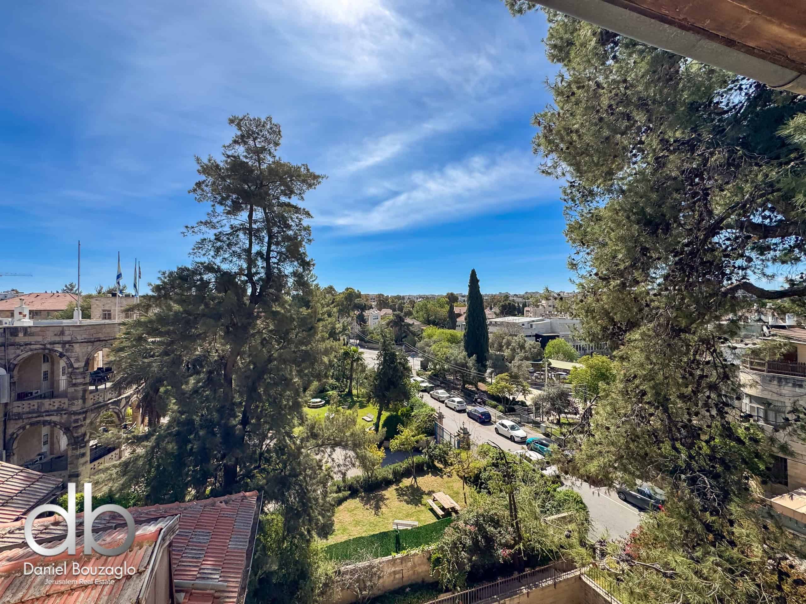 Sunny urban scene with tall trees, low-rise buildings, and parked cars under a clear blue sky.