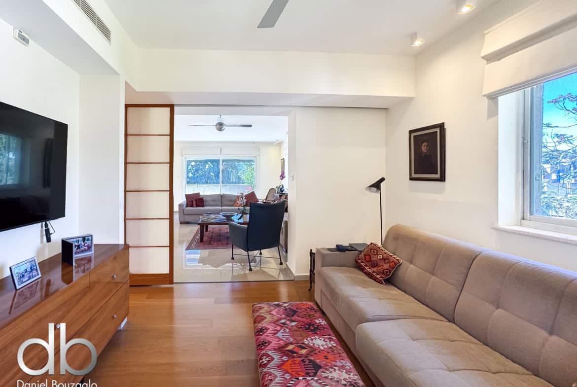 Bright, modern living room with a beige sectional sofa, a colorful patterned ottoman, a wooden sideboard, wall-mounted TV, ceiling fans, and a large window letting in natural light.