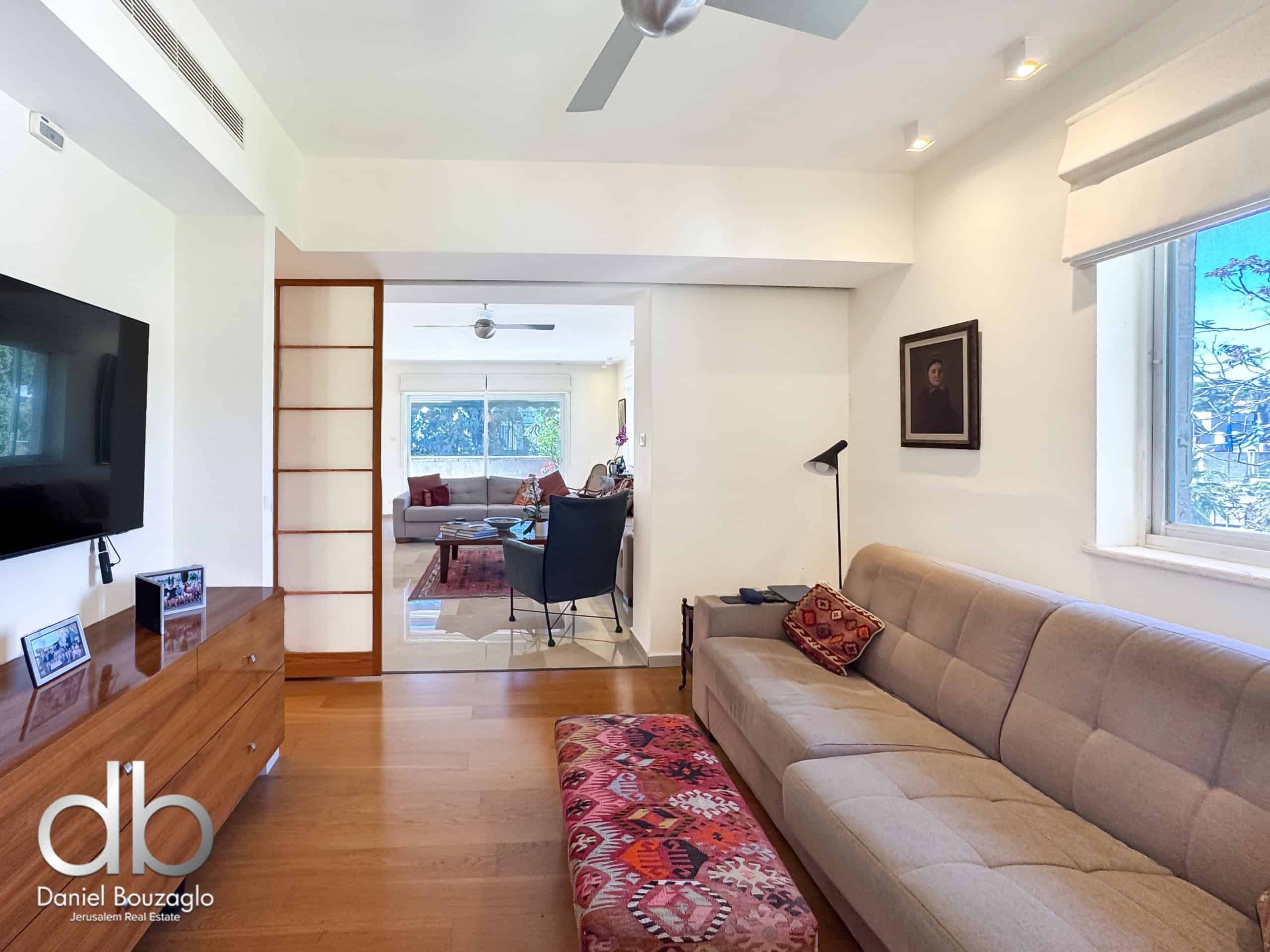 Bright, modern living room with a beige sectional sofa, a colorful patterned ottoman, a wooden sideboard, wall-mounted TV, ceiling fans, and a large window letting in natural light.