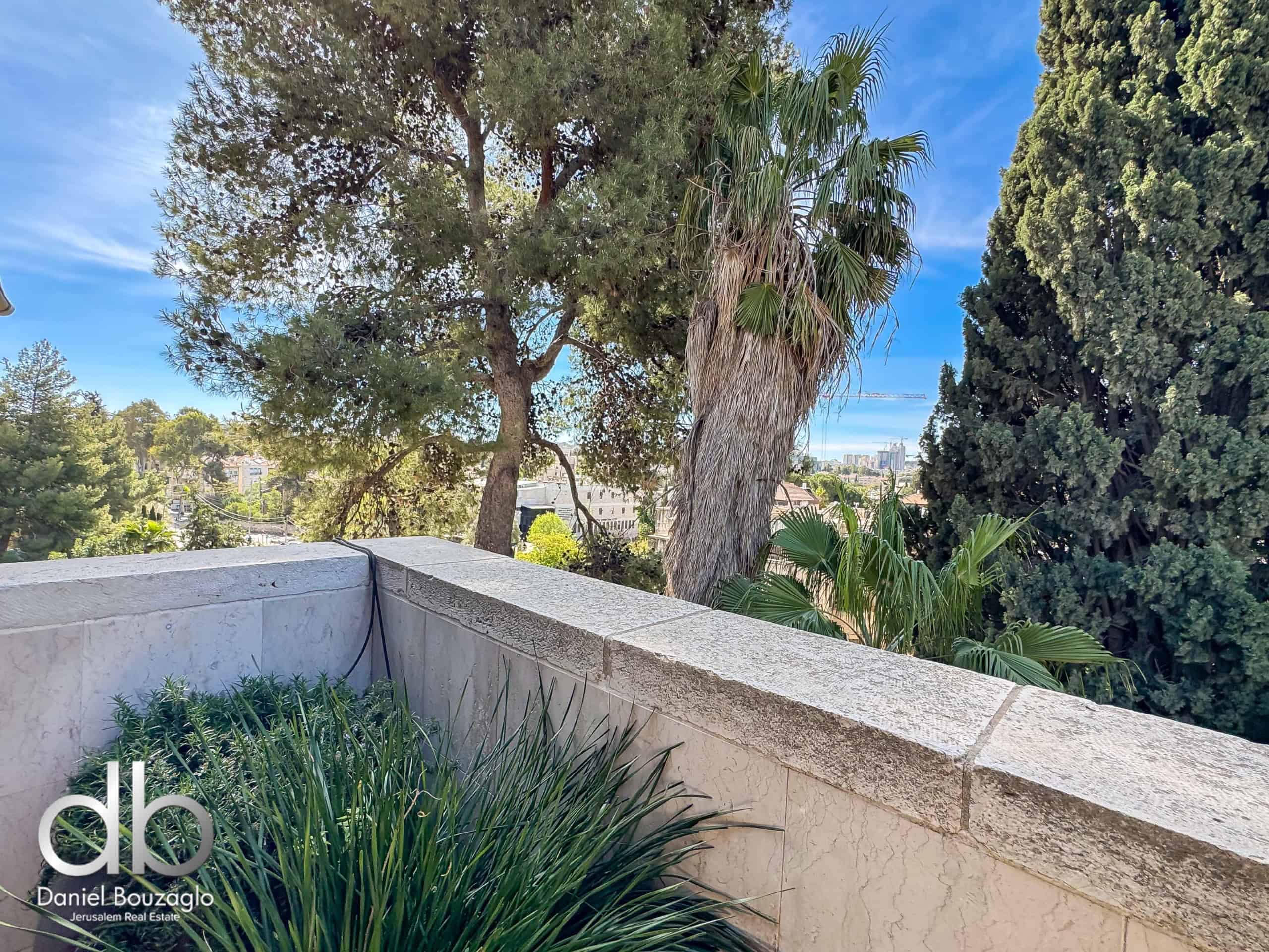 Terrace with a stone railing, lush green plants, and palm trees, overlooking a city skyline on a sunny blue-sky day; branding: Daniel Bouzaglio Jerusalem Real Estate.