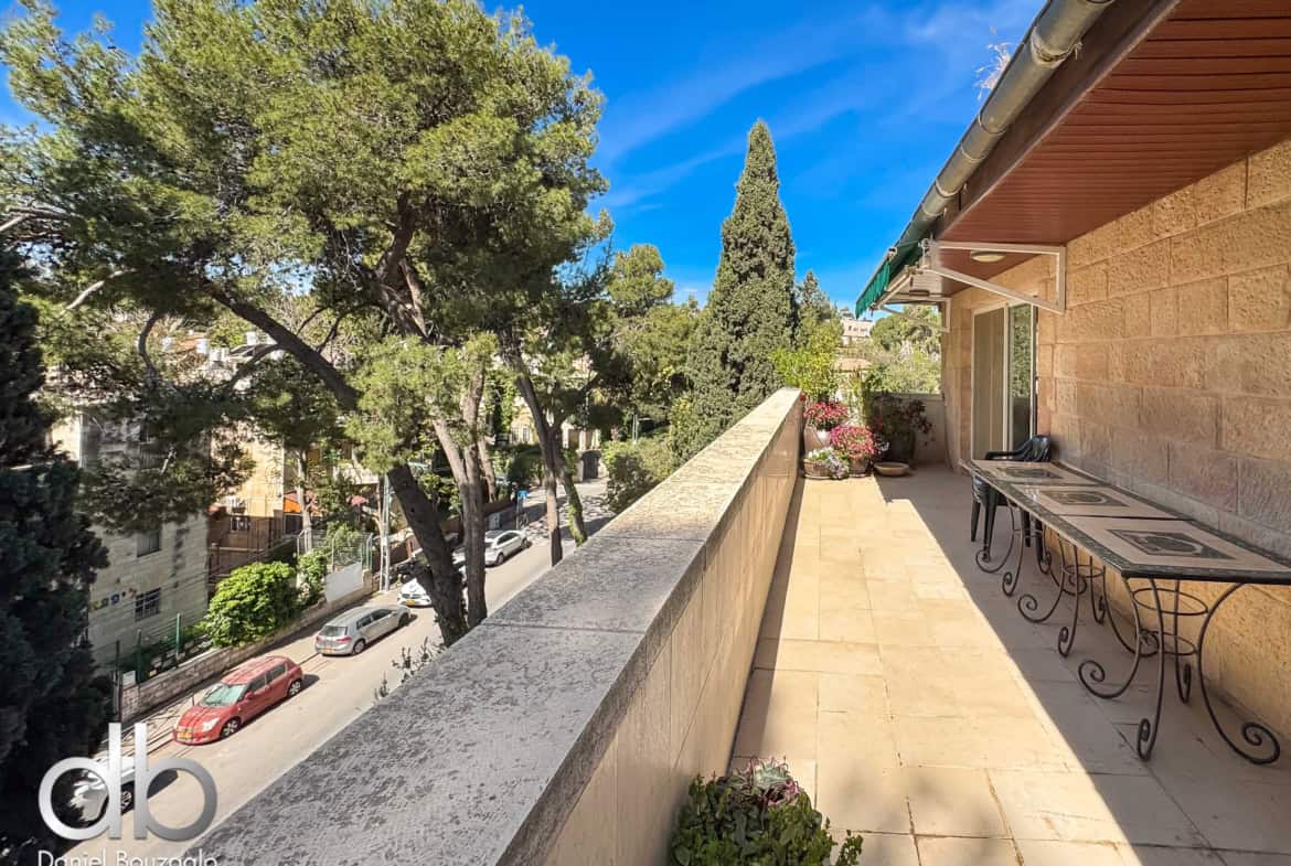 Balcony with tiled floor and wrought-iron tables, potted plants, overlooking a street lined with trees under a blue sky.
