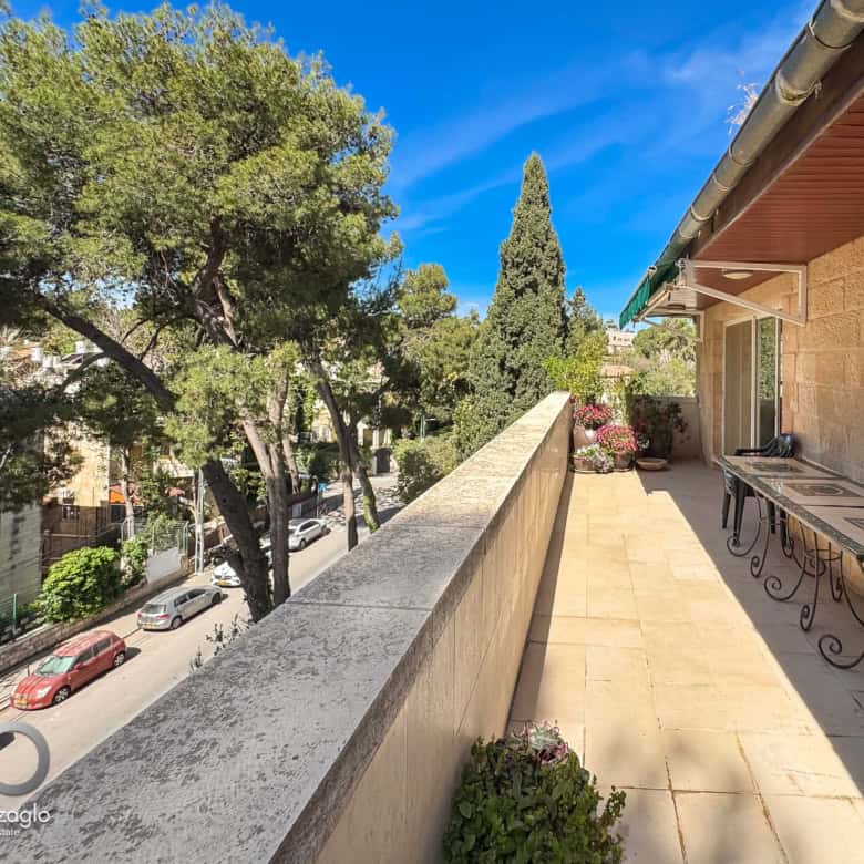 Balcony with wrought-iron tables and chairs overlooking a tree-lined street on a sunny day.