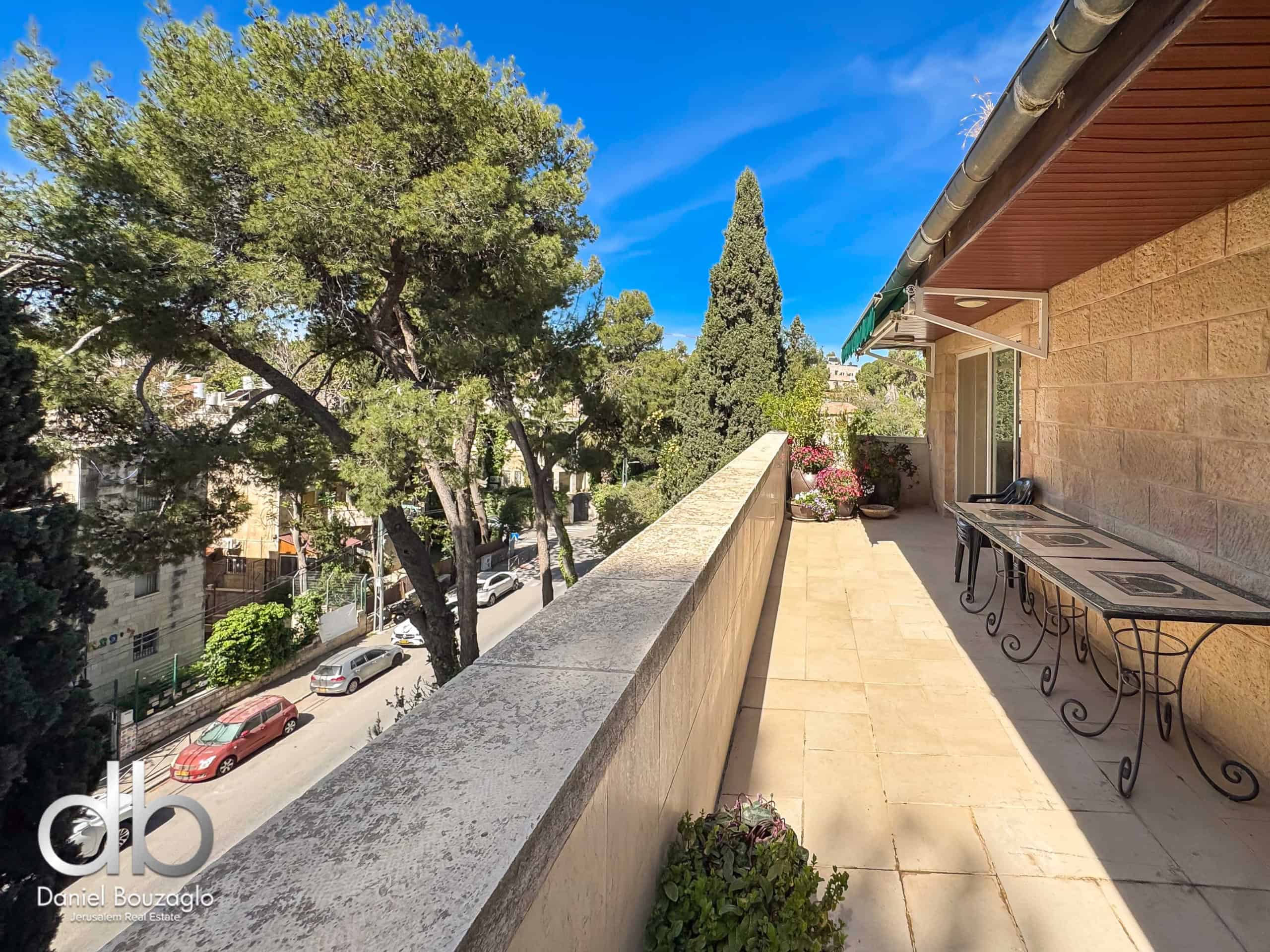 Balcony with tiled floor and wrought-iron tables, potted plants, overlooking a street lined with trees under a blue sky.