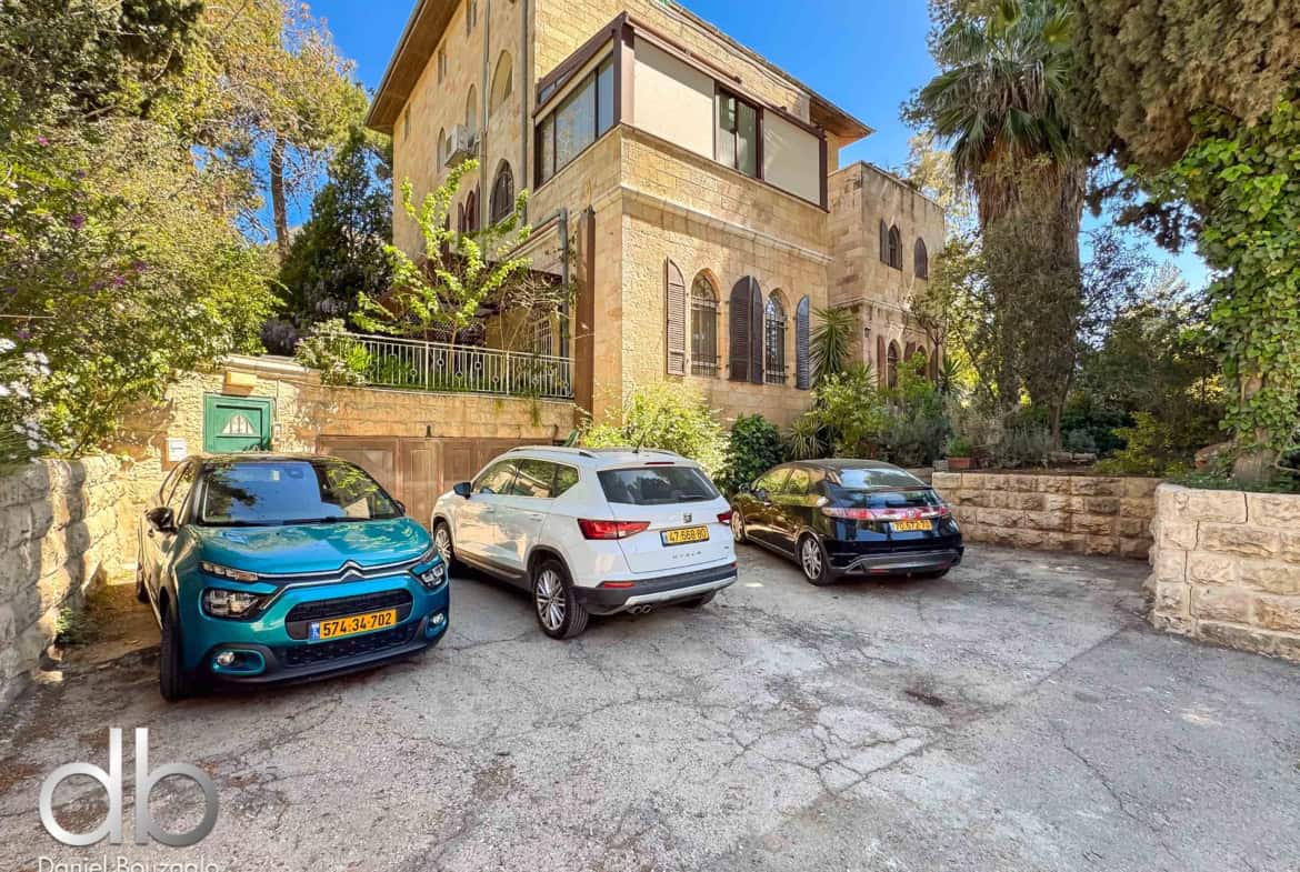 Stone two-story villa with arched windows, beige stone facade, and a small balcony, surrounded by trees, with several parked cars in a driveway.