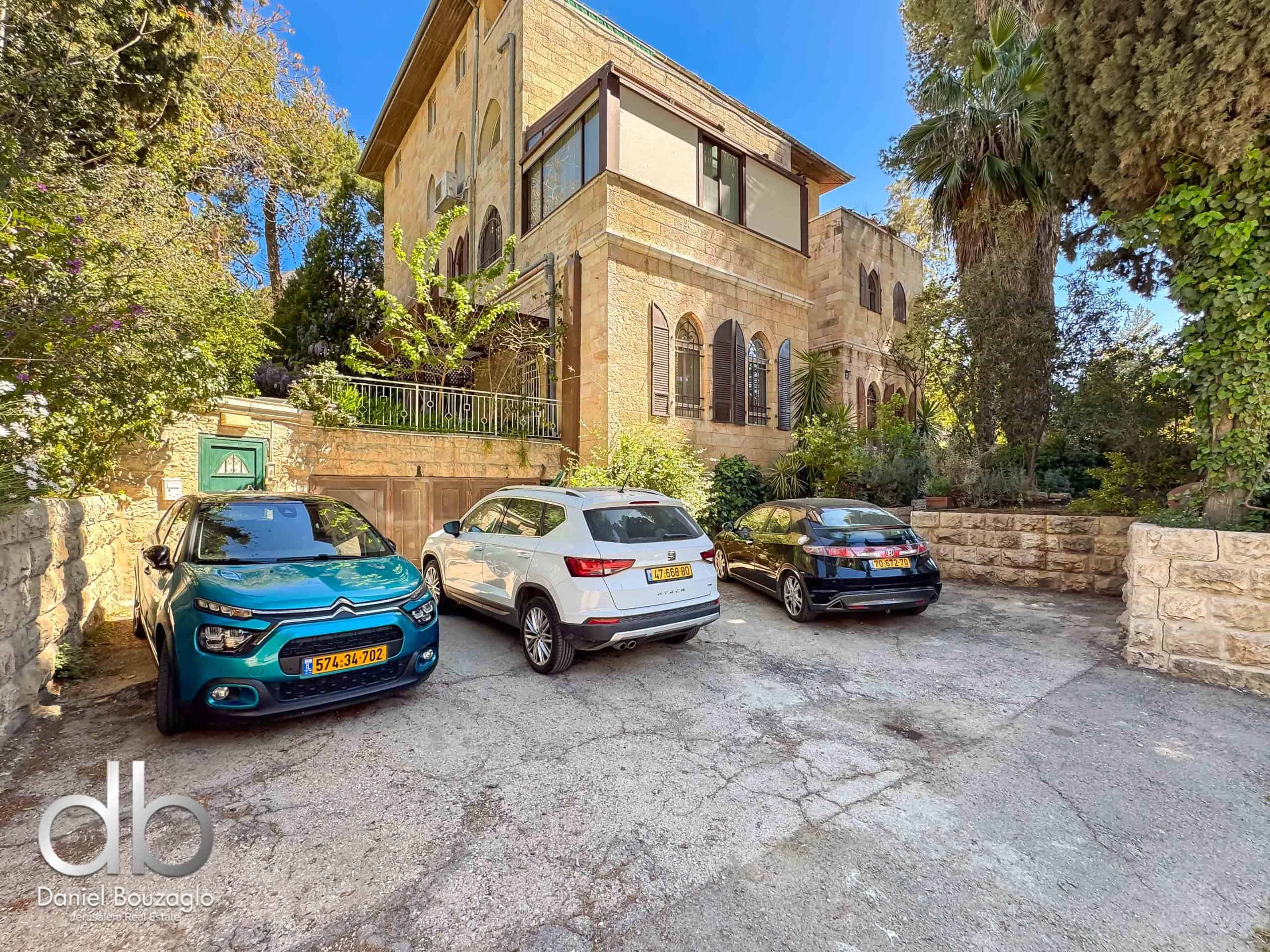 Stone two-story villa with arched windows, beige stone facade, and a small balcony, surrounded by trees, with several parked cars in a driveway.