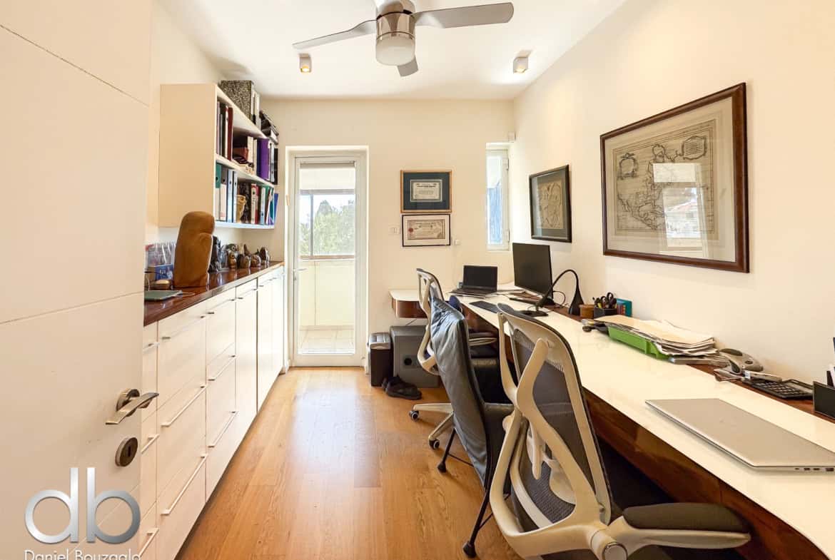 Bright home office with a long white desk on the right, ergonomic chairs, and built-in white cabinets along the left wall; ceiling fan overhead and framed maps on the walls.