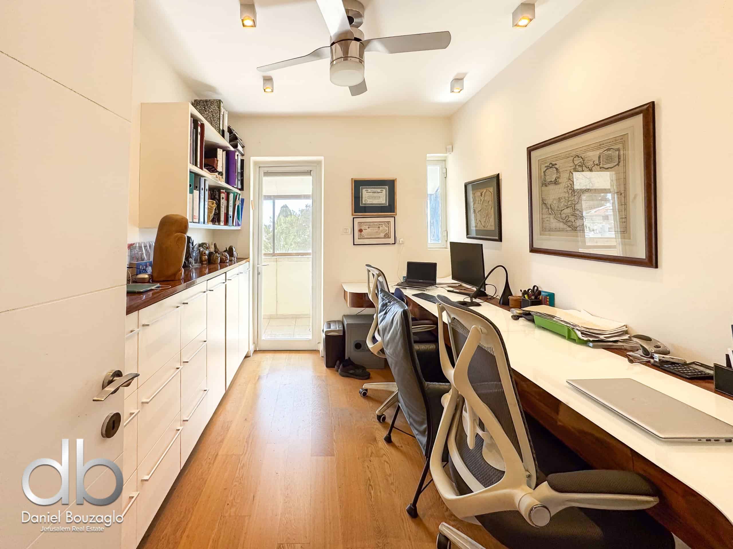 Bright home office with a long white desk on the right, ergonomic chairs, and built-in white cabinets along the left wall; ceiling fan overhead and framed maps on the walls.