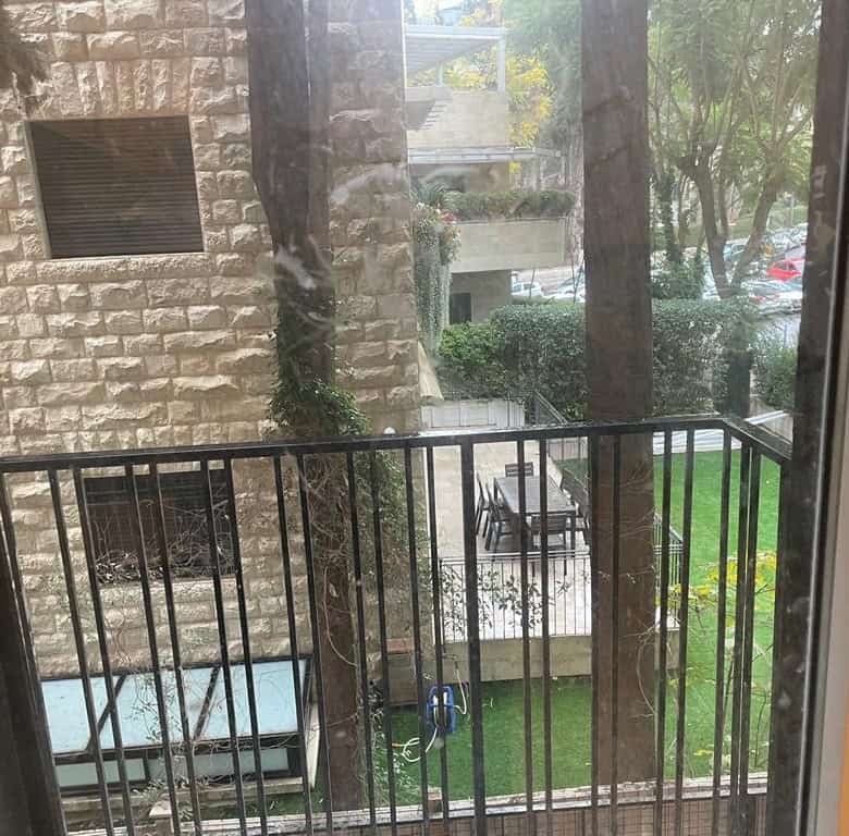View from a window onto a balcony with a black railing, a stone building wall, and an outdoor table with chairs on a patio, surrounded by trees and grass.