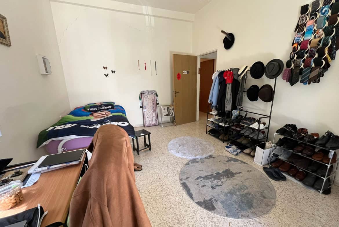 Bedside area with a blue-and-green blanket, left side, and a cluttered desk nearby in a small bedroom/closet space under a high ceiling.
