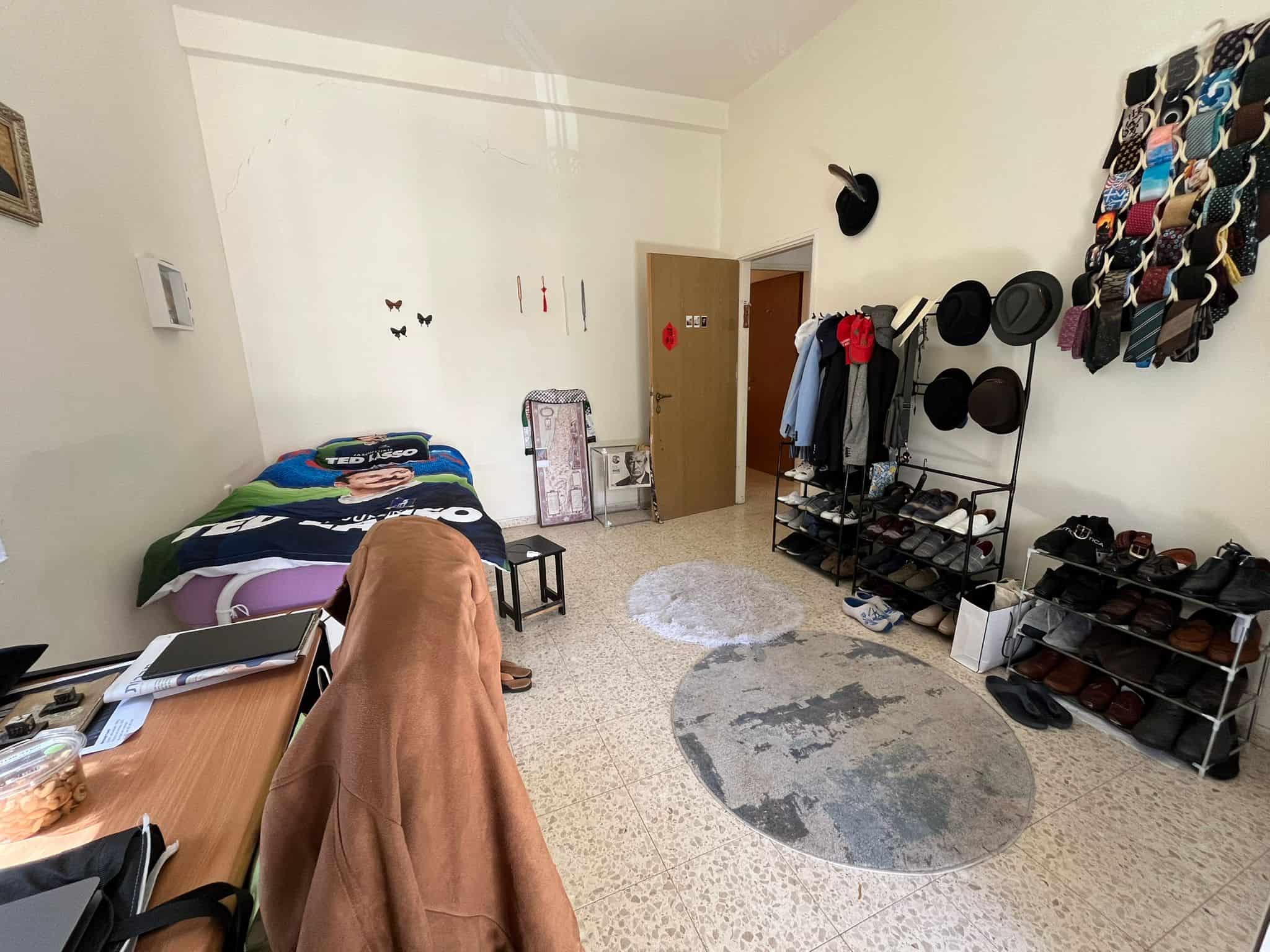 Bedside area with a blue-and-green blanket, left side, and a cluttered desk nearby in a small bedroom/closet space under a high ceiling.