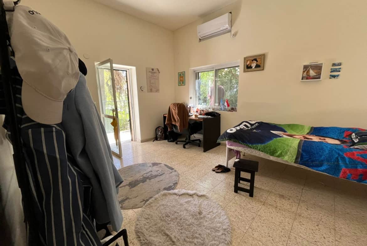 Bedroom scene: a single bed with a colorful quilt on the right, desk and chair under a window, and an air conditioner above.