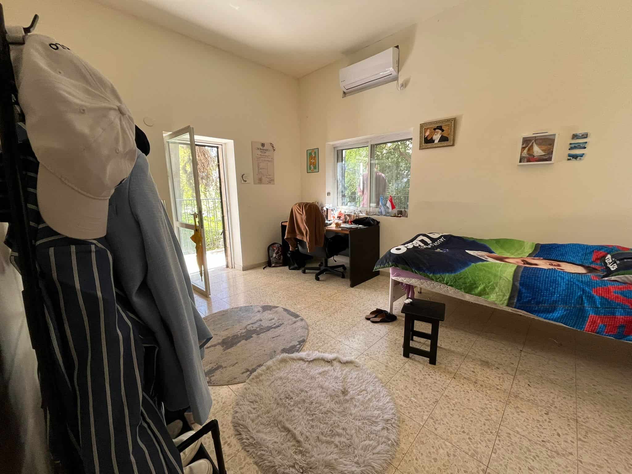 Bedroom scene: a single bed with a colorful quilt on the right, desk and chair under a window, and an air conditioner above.