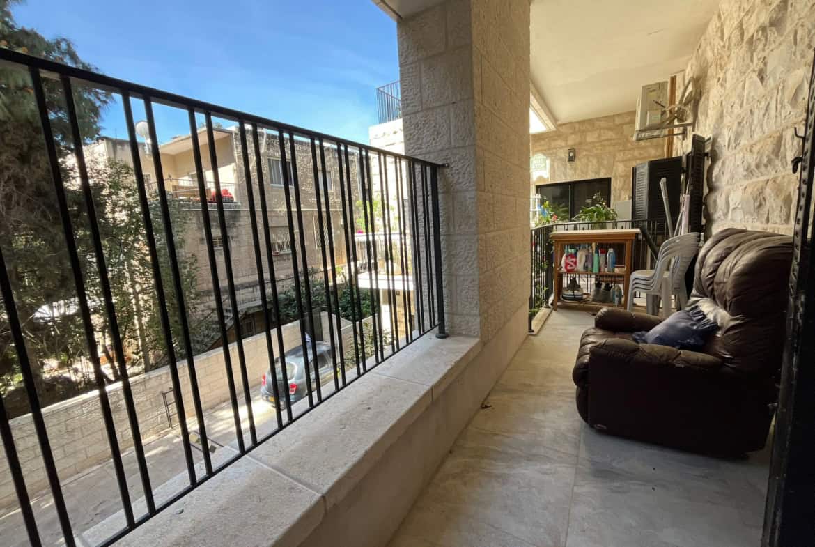 Covered apartment balcony with a brown leather sofa, plastic chairs, and cleaning supplies on a small table against stone walls and a metal railing nearby.