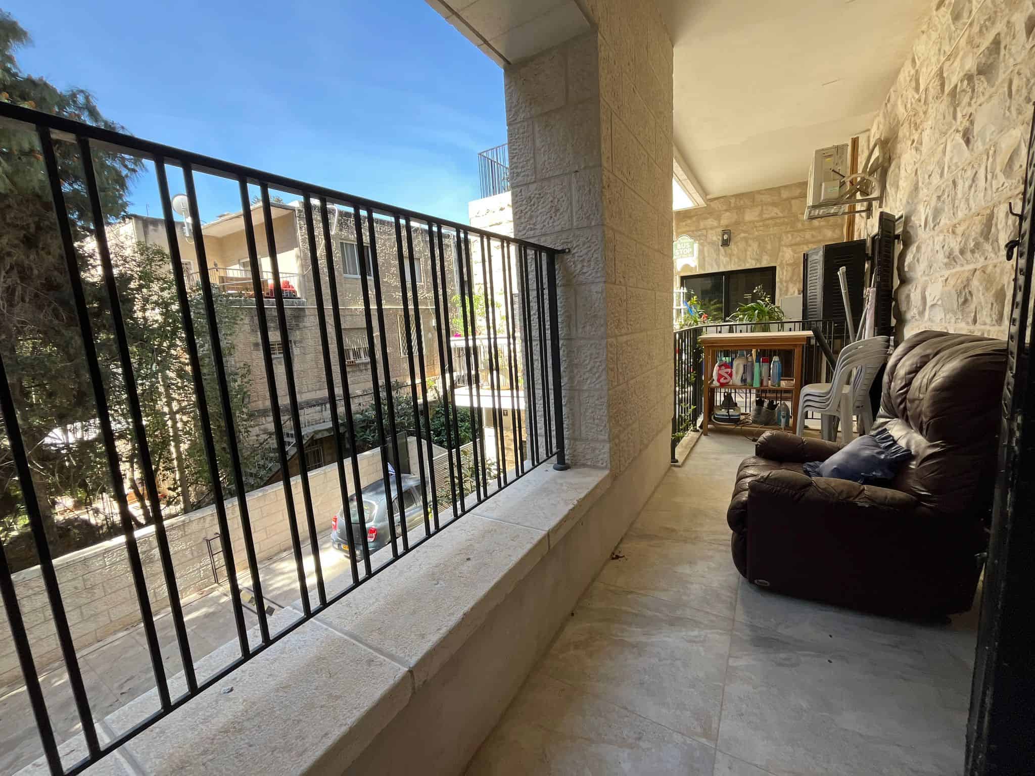 Covered apartment balcony with a brown leather sofa, plastic chairs, and cleaning supplies on a small table against stone walls and a metal railing nearby.