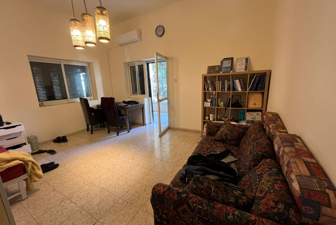 Living room with a patterned sofa on the right, tall bookshelf, and hanging pendant lights; door to balcony visible in background.