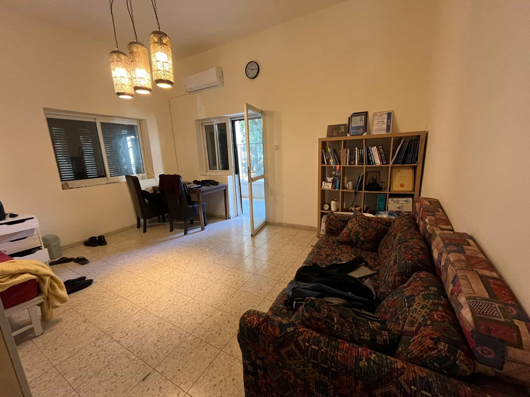 Living room with a patterned sofa on the right, tall bookshelf, and hanging pendant lights; door to balcony visible in background.