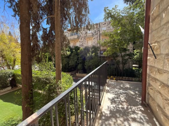 Balcony with metal railing overlooks a courtyard and stone apartment building, with green trees and blue sky in bright sunlight.