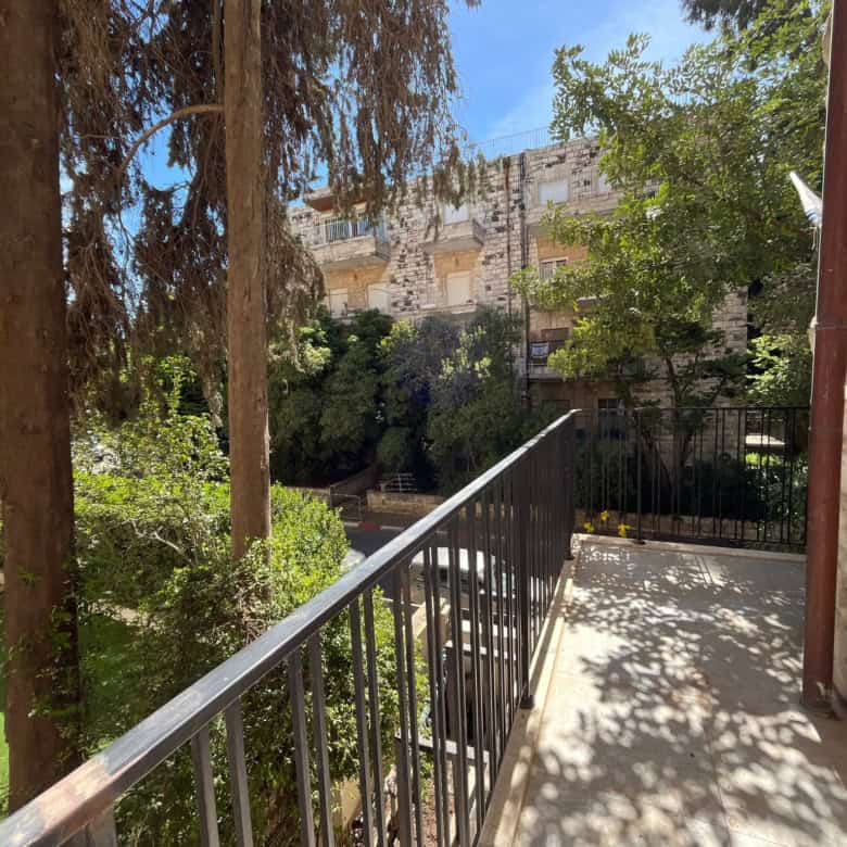 Balcony with metal railing overlooks a courtyard and stone apartment building, with green trees and blue sky in bright sunlight.