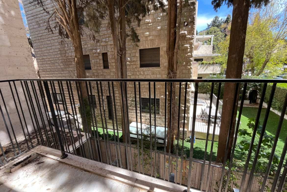 Balcony with a metal railing overlooking a courtyard and beige stone building with trees nearby. Carved shadows on the floor.