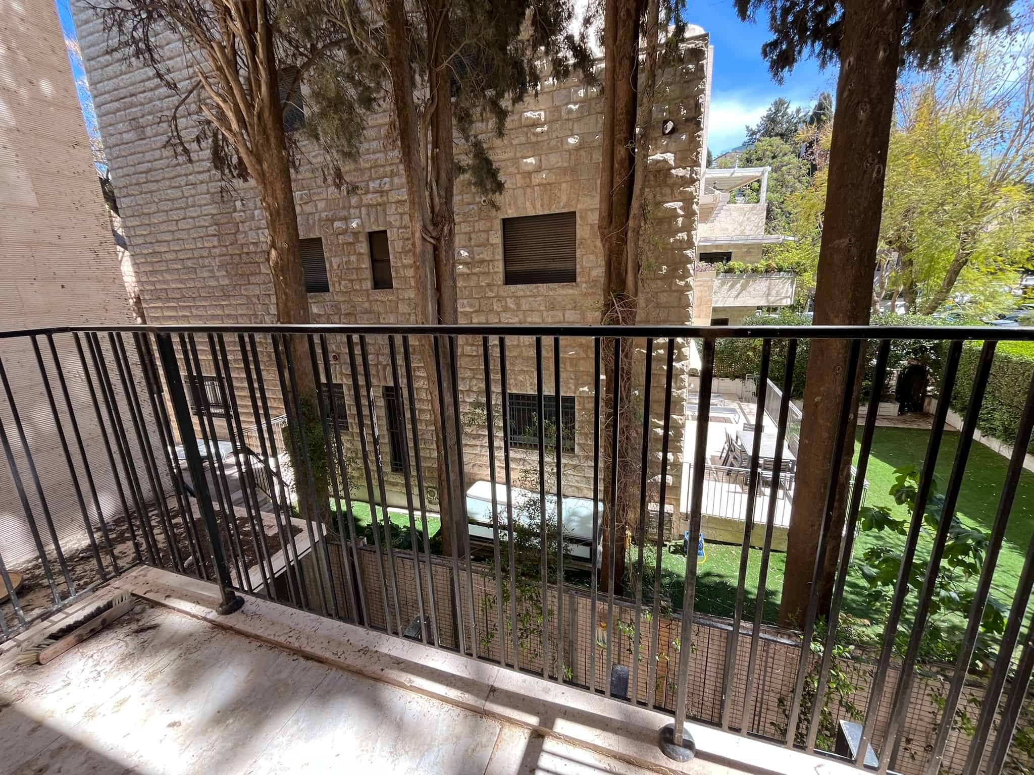 Balcony with a metal railing overlooking a courtyard and beige stone building with trees nearby. Carved shadows on the floor.