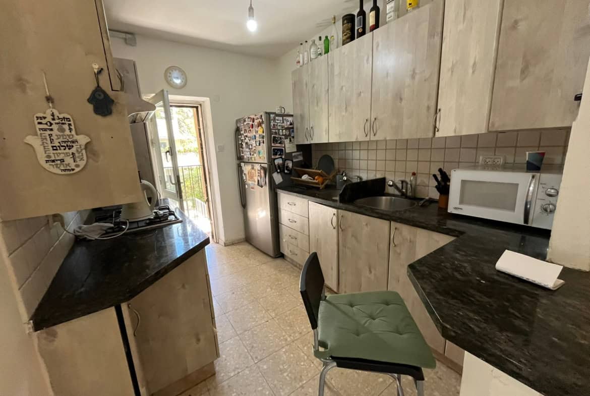 Galley kitchen with light wood cabinets, black countertops, and a fridge covered in magnets along the left wall; microwave on the right counter.