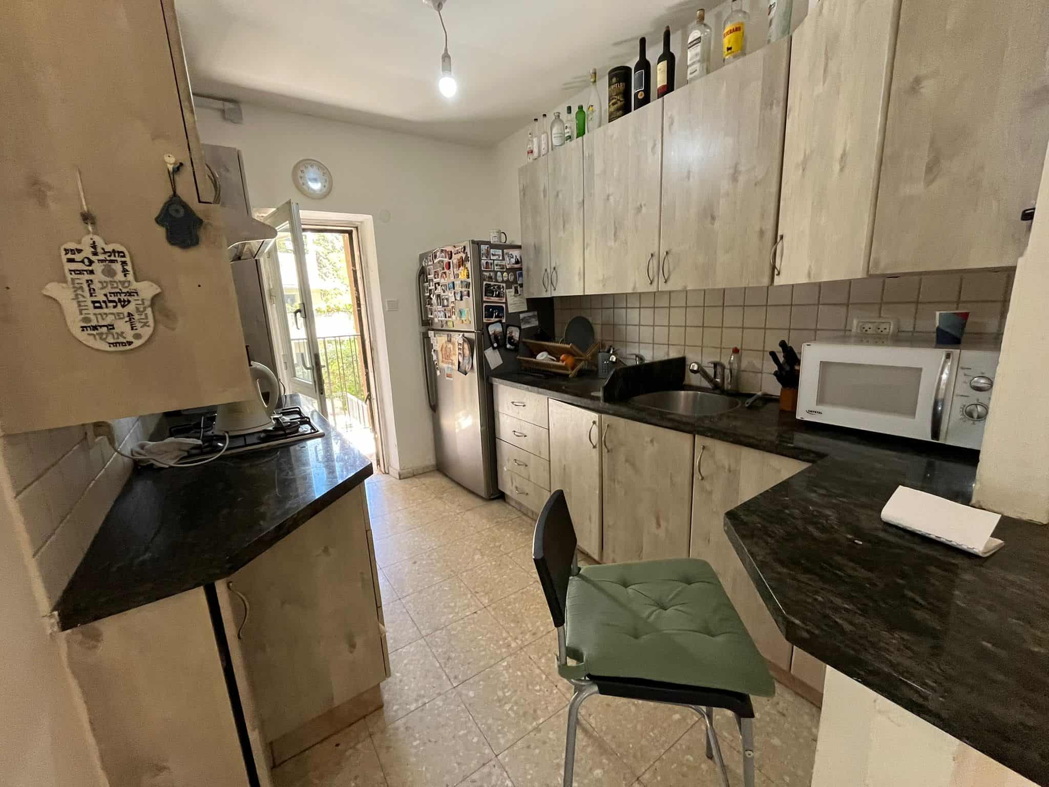 Galley kitchen with light wood cabinets, black countertops, and a fridge covered in magnets along the left wall; microwave on the right counter.