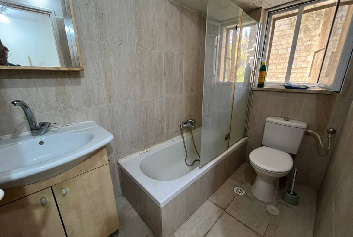 Bathroom with a sink on the left, tiled walls, a glass shower screen, a bathtub, and a toilet by a window; a cleaning bottle sits on the sill.
