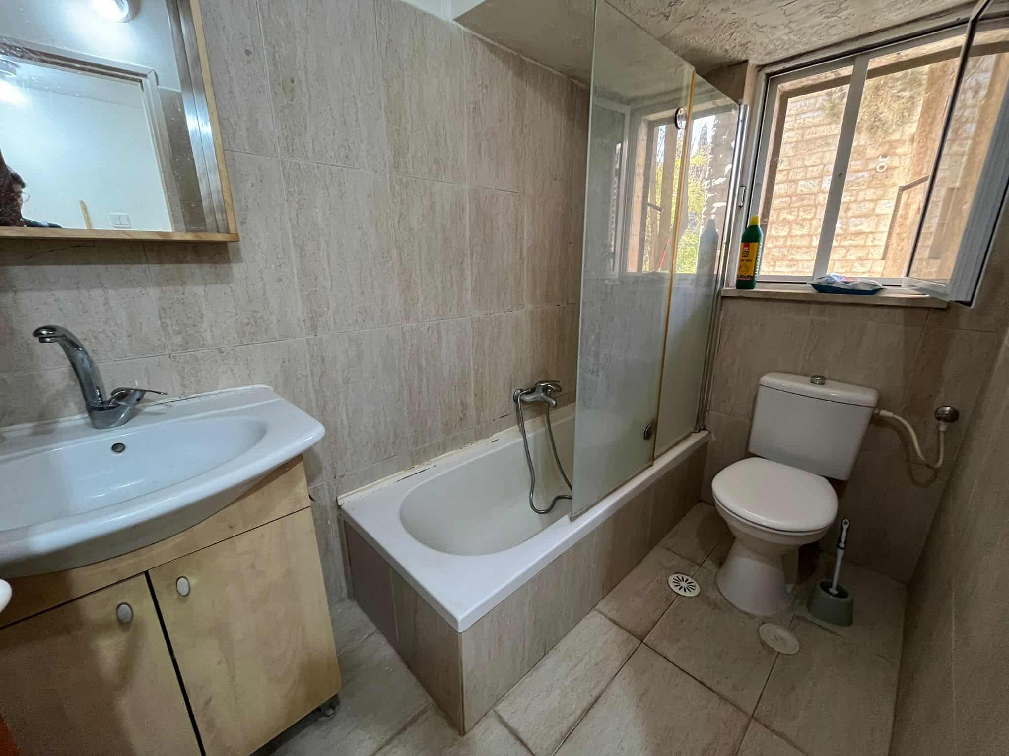 Bathroom with a sink on the left, tiled walls, a glass shower screen, a bathtub, and a toilet by a window; a cleaning bottle sits on the sill.