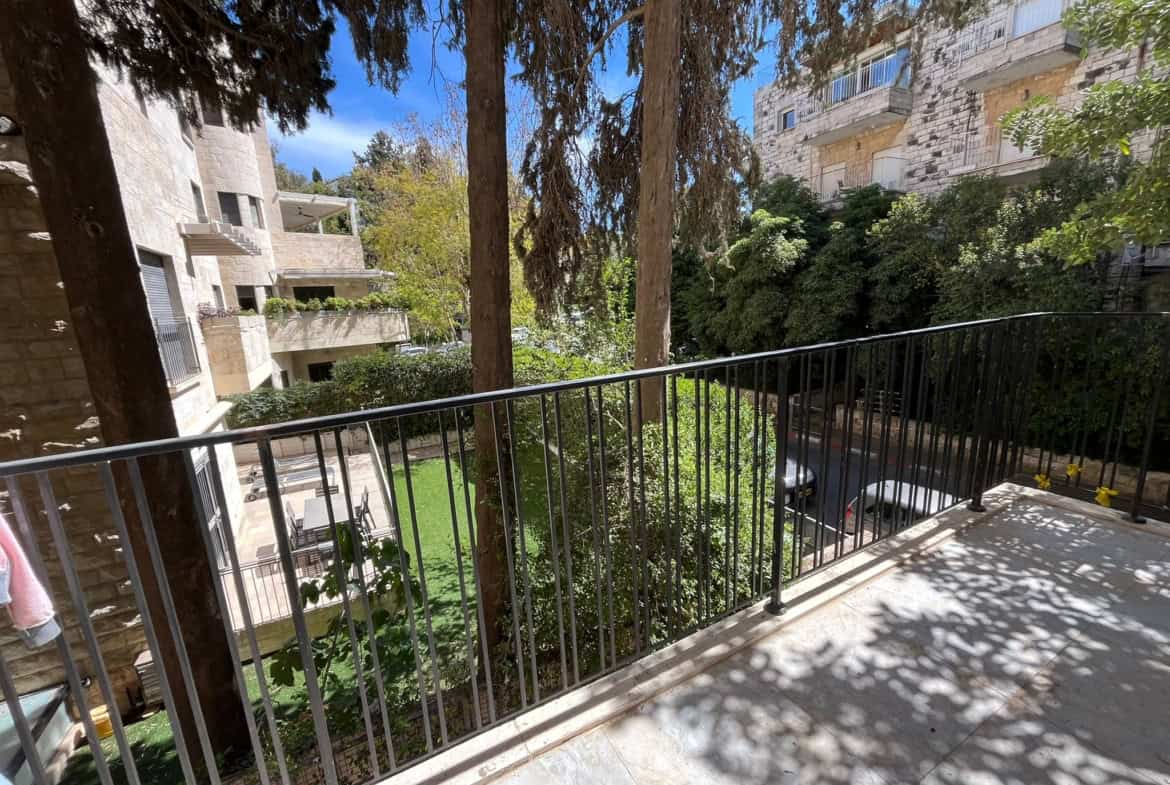 Balcony railing with shadows overlooking a courtyard, flanked by stone apartment buildings and tall trees.
